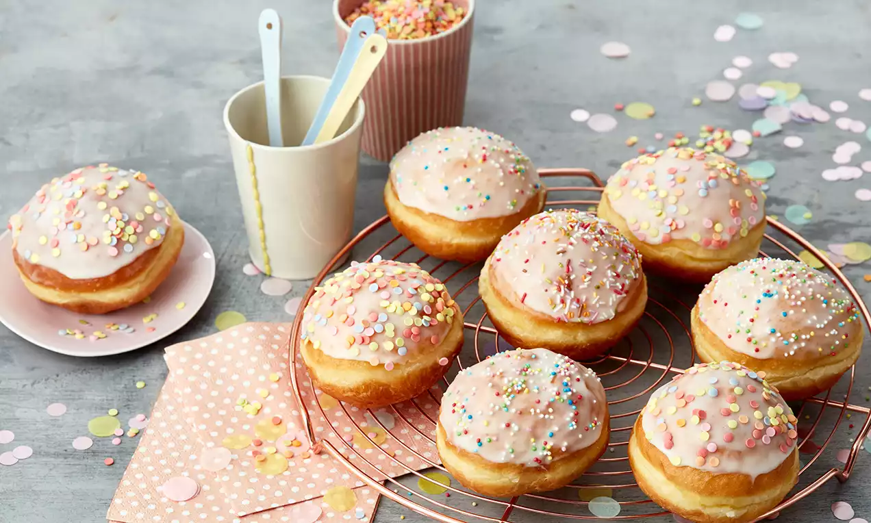 Beignets dorés et moelleux glacés de rose pâle, parsemés de confettis colorés sur une grille métallique entourée de décorations pastel