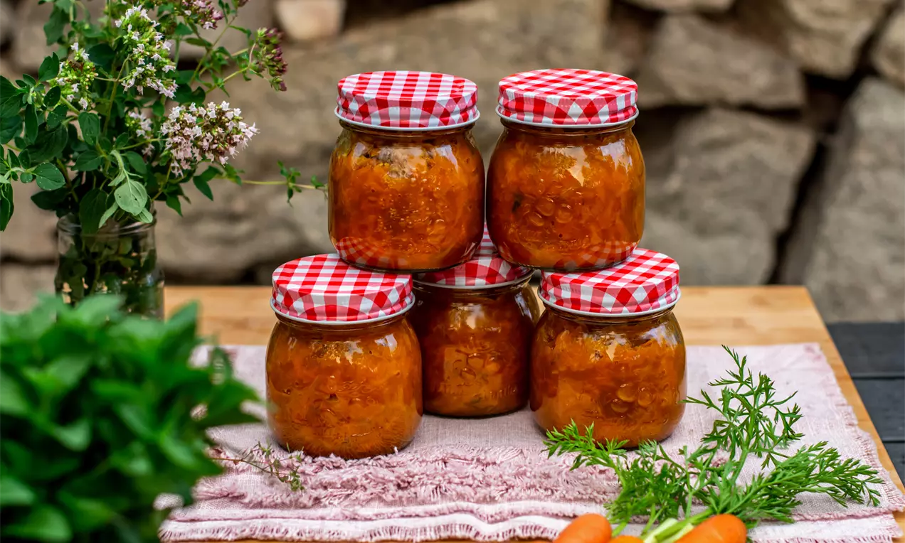 Пет стъклени буркана с ярко оранжево чътни от моркови и червено-бели карирани капачки върху розова покривка, заобиколени от зелени билки и пресни моркови