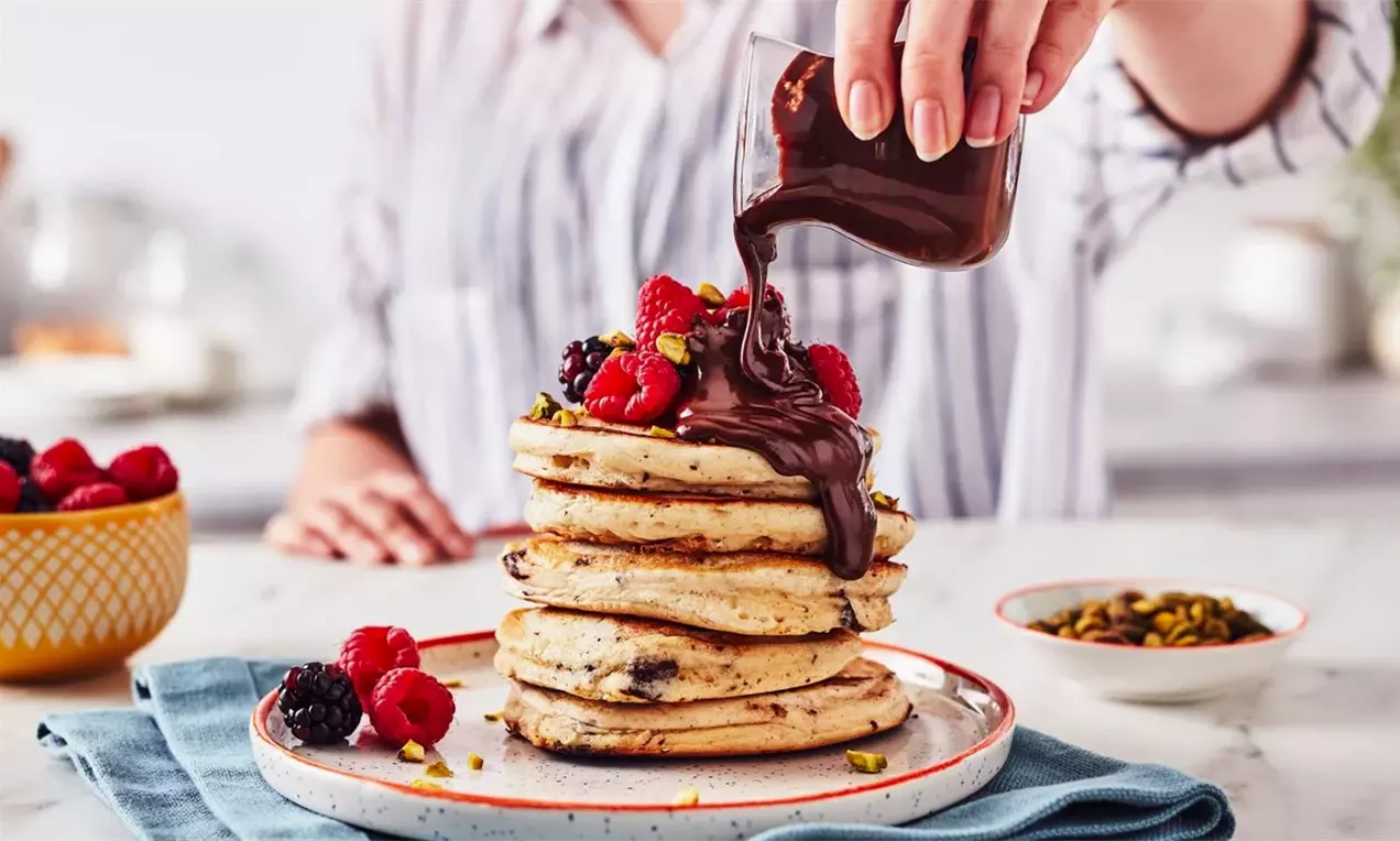 Висока купчина златисти палачинки с шоколадова глазура, гарнирани с пресни ягоди и шамфъстък върху бяла чиния с синя салфетка