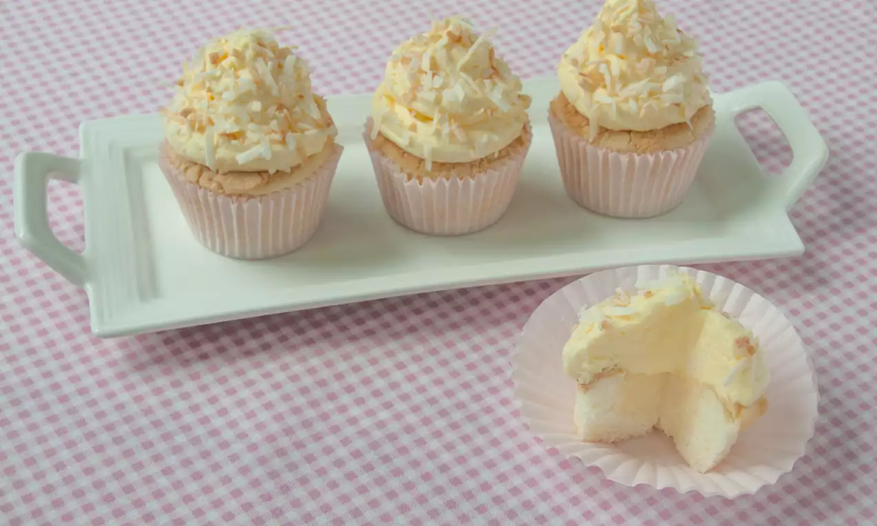 Cupcakes moelleux dans des caissettes rose pâle, ornés de crème blanche et de copeaux de noix de coco sur un plateau rectangulaire blanc
