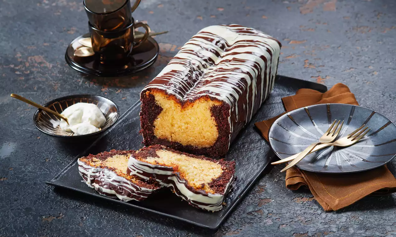Dunkelbrauner Kastenkuchen mit heller Füllung, umhüllt von weißer und dunkler Schokoladenglasur, serviert auf schwarzer Platte mit Geschirr