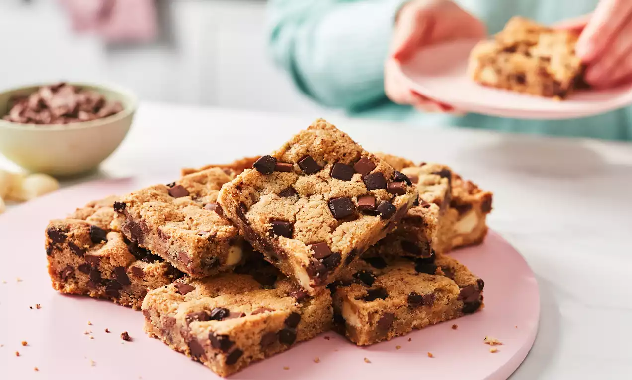 Square cookie bars stacked on a round pastel pink plate, golden brown with dark chocolate chunks, set on a white countertop