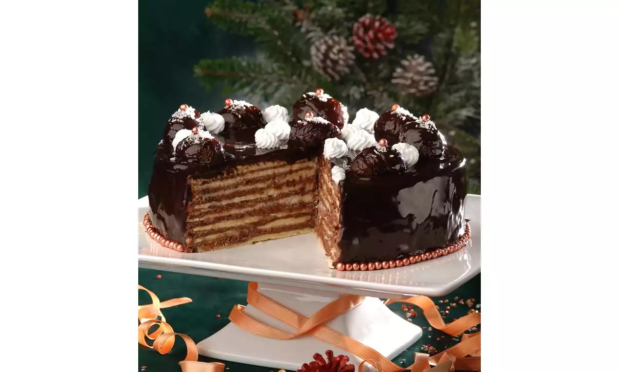 Kat kat ince şeritli kestaneli pasta, parlak çikolata kaplaması, beyaz krema rozetleri ve çam dalları süslemeli bir servis tabağında yer alıyor