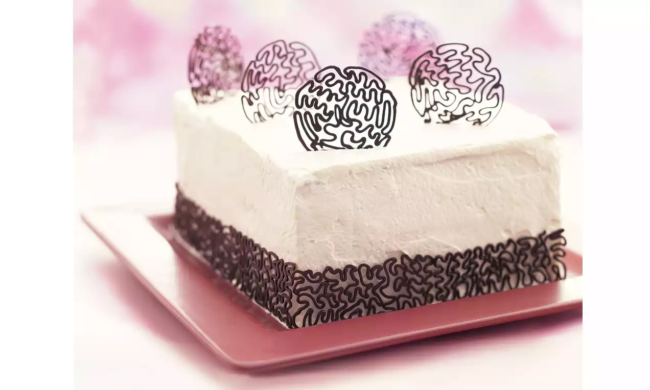 Weiße rechteckige Torte mit glatter Sahnecreme, verziert mit dunklen filigranen Ornamenten und rundem Schokolatendekor auf rosa Teller