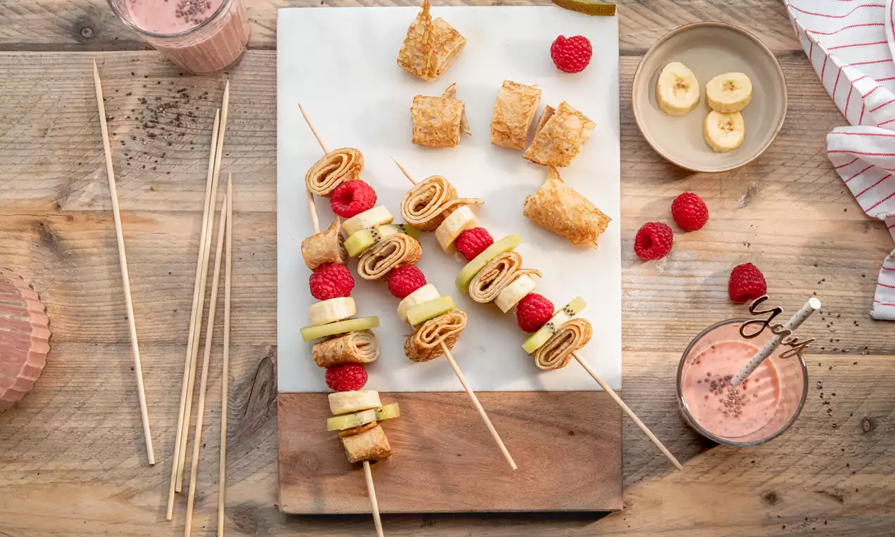 Brochettes de crêpes roulées dorées, intercalées de tranches de banane, framboises rouges et morceaux de kiwi vert sur un plateau en bois et marbre