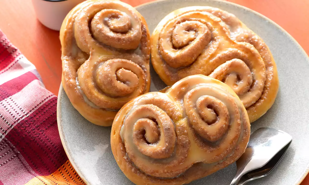 Fahéjas csigák aranyszínű spiráljai cukormázzal borítva, puhán és omlósan, szürke tányéron kiskanállal, mellette textil szalvéta