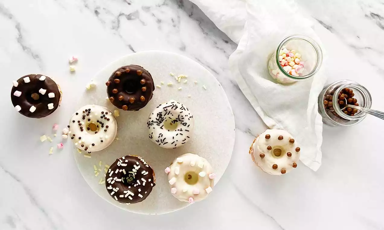 Mini Donuts in verschiedenen Glasuren mit bunten Streuseln und Dekoren auf einer weißen Platte, arrangiert auf marmoriertem Untergrund