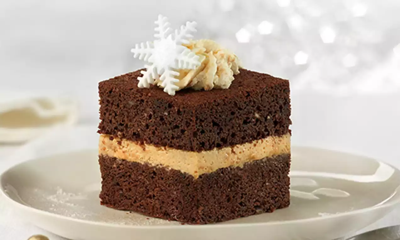Cubo de bizcocho de chocolate oscuro con capa central de mousse de turrón beige, decorado con crema y copo de nieve blanco sobre plato
