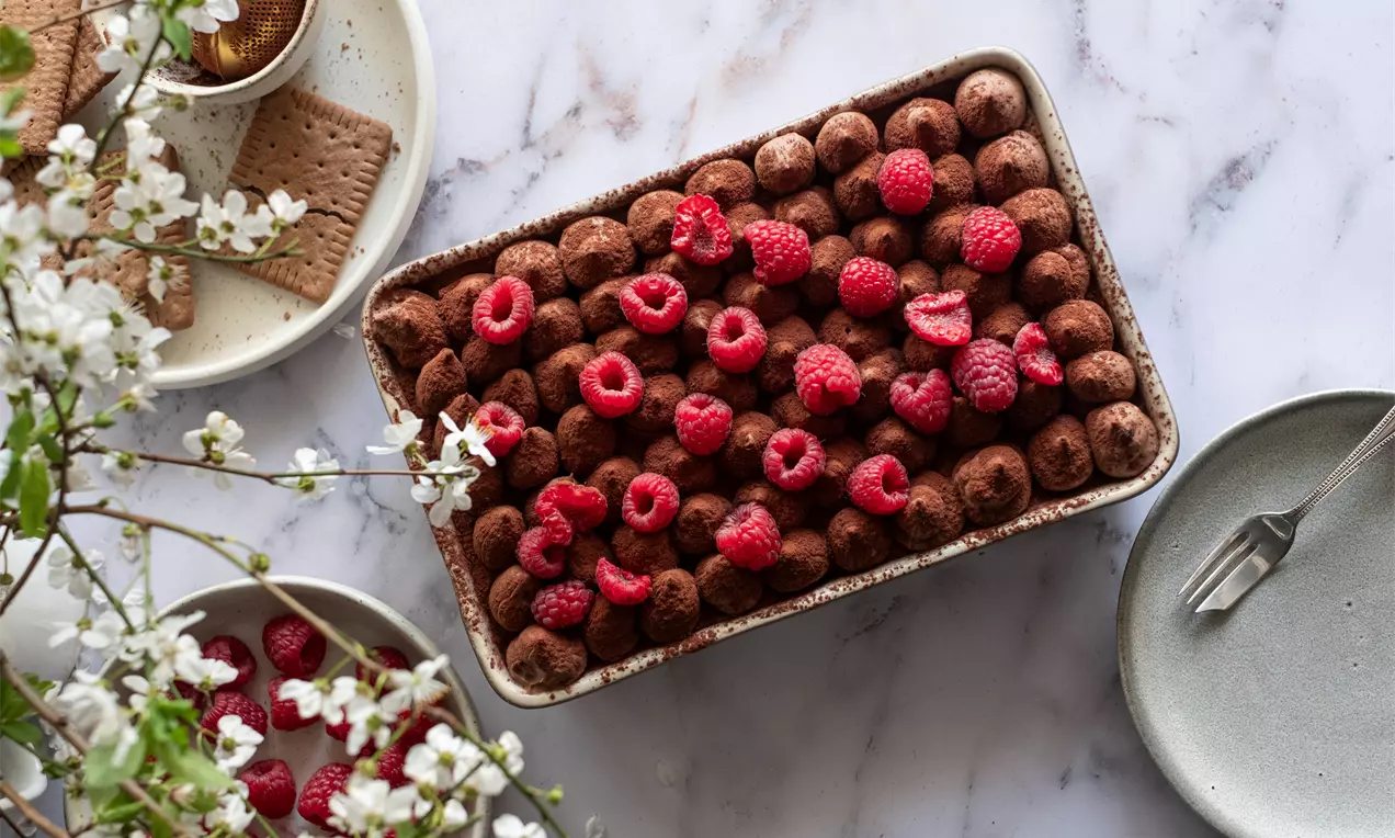 Шоколадова торта с мека, тъмна текстура, покрита с какаови топчета и свежи яркочервени малини в правоъгълна форма върху светъл фон