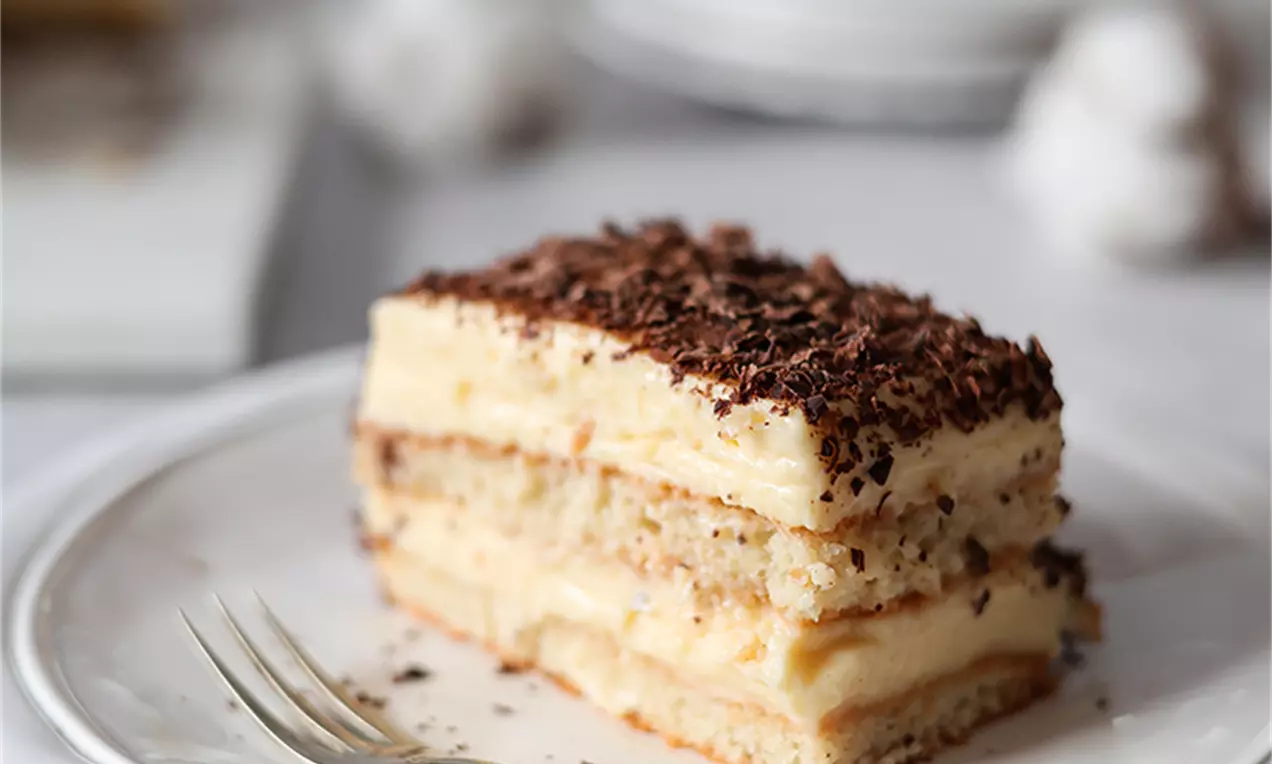 Деликатно парче бадемова торта с пластове светъл крем и бисквитен блат, поръсено с шоколадови стърготини върху бяла чиния