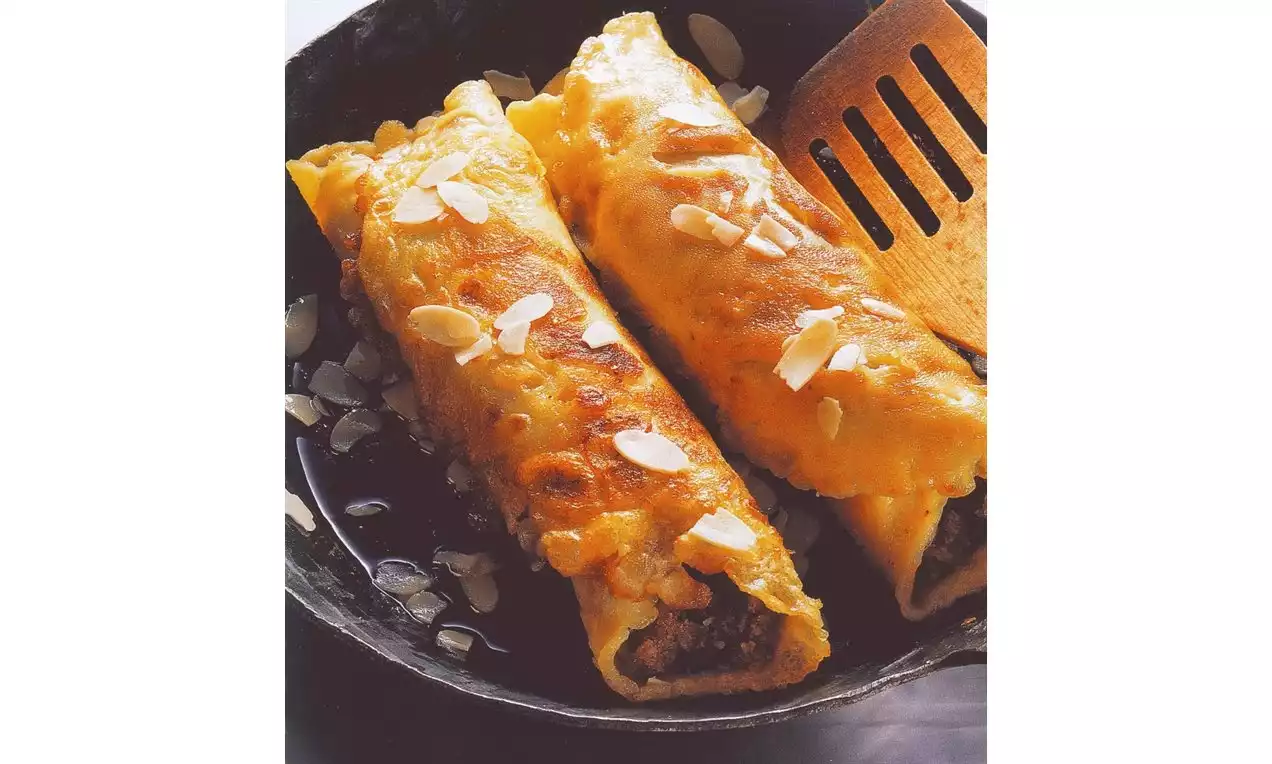 Crêpes dorées roulées garnies de noix, parsemées de fines lamelles d'amandes, servies dans une poêle noire avec une spatule en bois