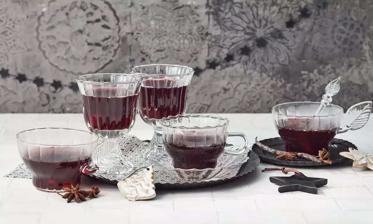 Dunkelrote Glühweinfüllung in verschieden geformten gläsernen Tassen auf silbernen und schwarzen Unterlagen mit Sternanis und Deko