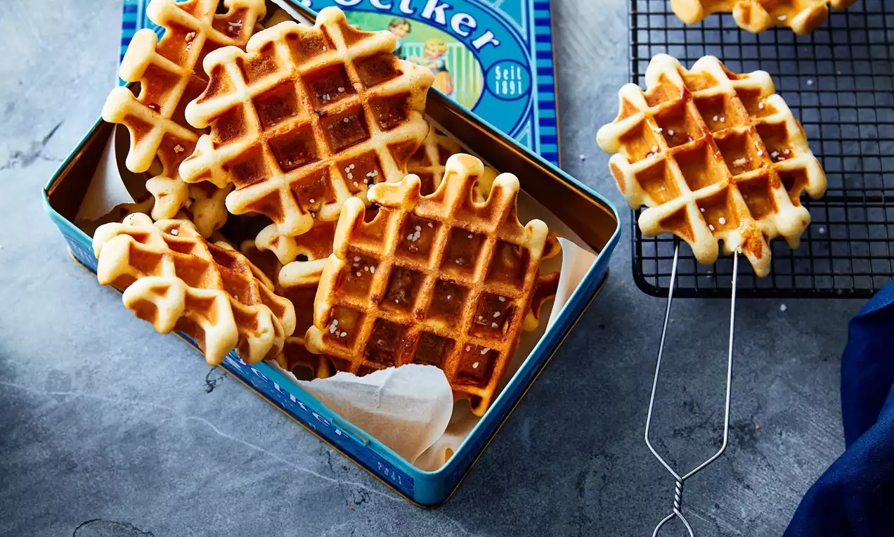 Luikse wafels met een goudbruine kleur en stevige textuur in een blauwe metalen doos, naast een zwarte koelrek op een grijze ondergrond