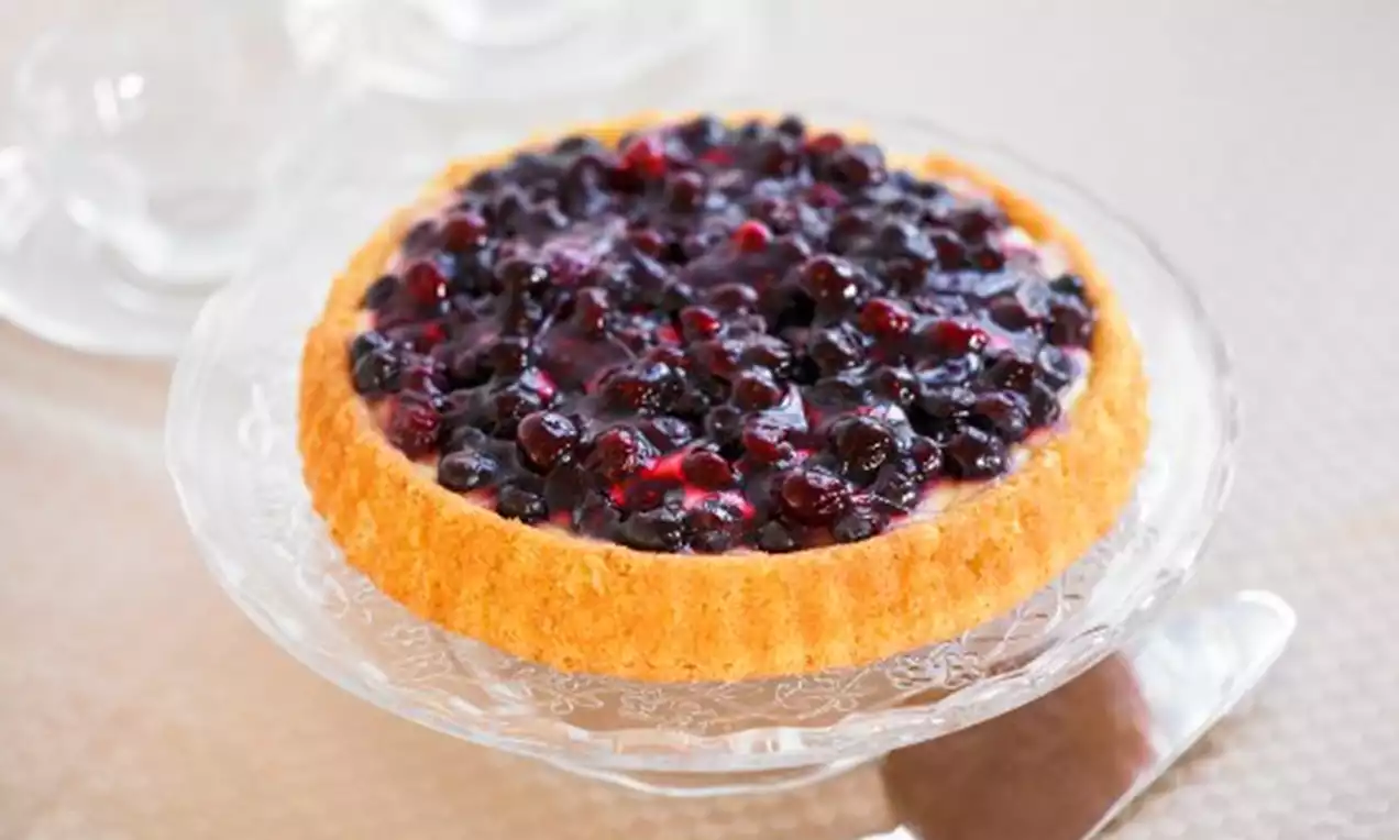 Rund mördegskaka med gyllene botten täckt av glansig blåbärsfyllning, serverad på en glastallrik med en silverspade bredvid
