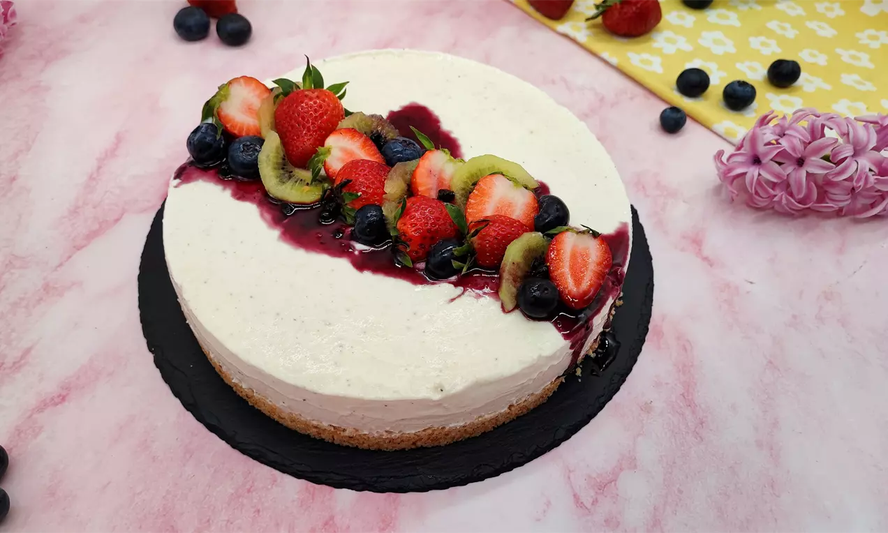 Кръгъл чийзкейк с кремообразнa повърхност и златисто бисквитено дъно, увенчан със свежи ягоди, киви и боровинки върху лилав плодов сос
