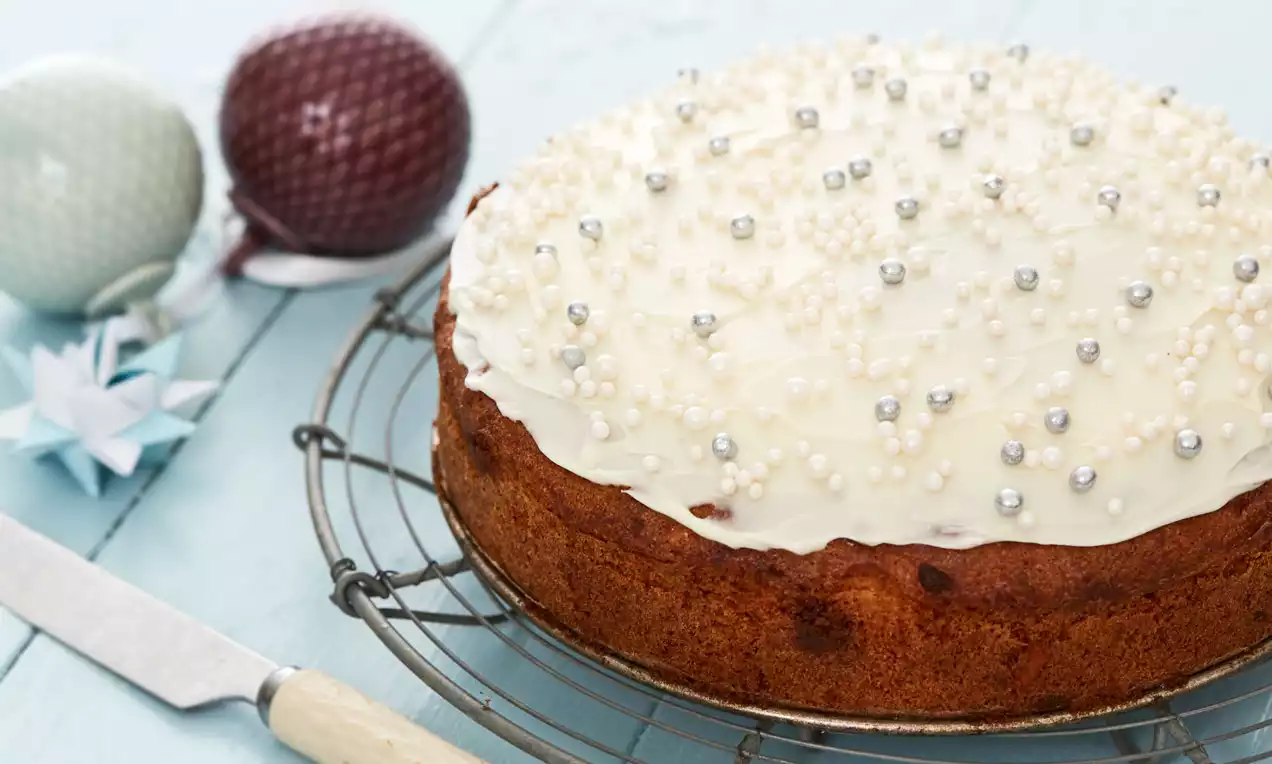 Rund vinterkake med gyllenbrun kant, hvit glasur toppet med små perler og sølvkuler, plassert på en metallrist mot blå bakgrunn