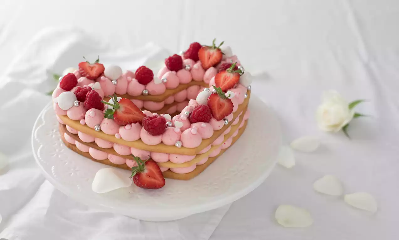 Hjärfformad tårta med lager av gyllene mördeg och rosa frosting, dekorerad med röda hallon, jordgubbshalvor och vita sockerpärlor på vit tårtbricka