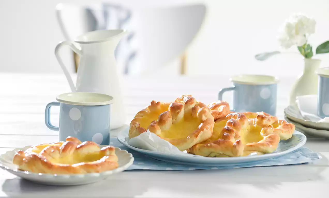 Goldbraune Puddingbrezeln mit glänzender Oberfläche und gelber Füllung auf einem weißen Teller, arrangiert neben blauen Tassen mit Punkten
