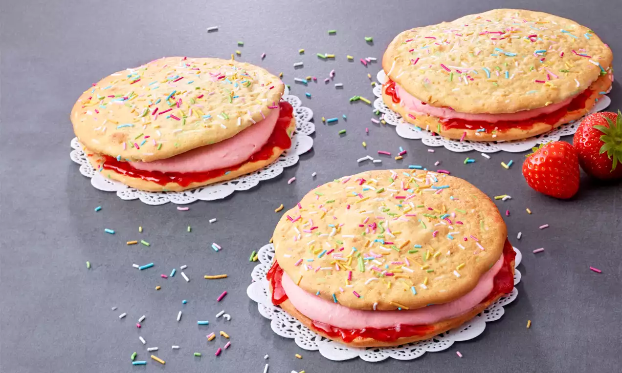 Runde Omelettes mit hellgelbem Teig, gefüllt mit roter Erdbeermarmelade und rosa Creme, bestreut mit bunten Streuseln auf Spitzendeckchen