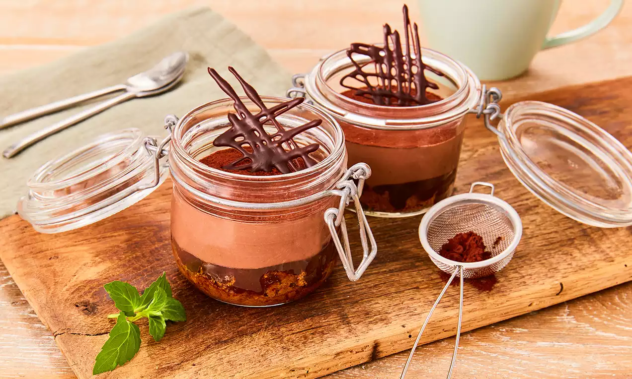 Veganes Schokoladen-Tiramisu in gläsernen Weckgläsern mit geschichteter Creme und dunkler Schokogarnitur auf Holzbrett mit Sieb und Minze