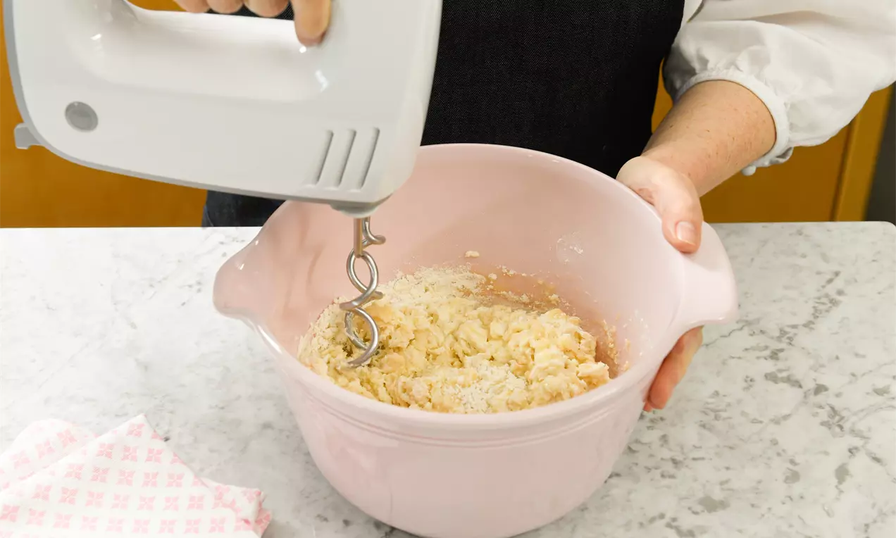 Светложълто тесто с едра, ронлива текстура се смесва с миксер в бледорозова купа върху мраморен плот с розово бяло каре кърпа