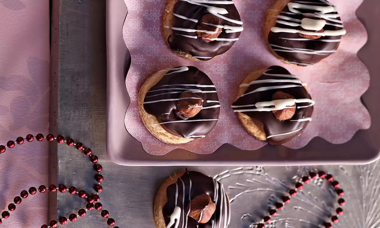 Kulaté oříškové dortíky na růžovém podnose, zdobené lesklou tmavou čokoládou, bílými proužky a celými lískovými oříšky