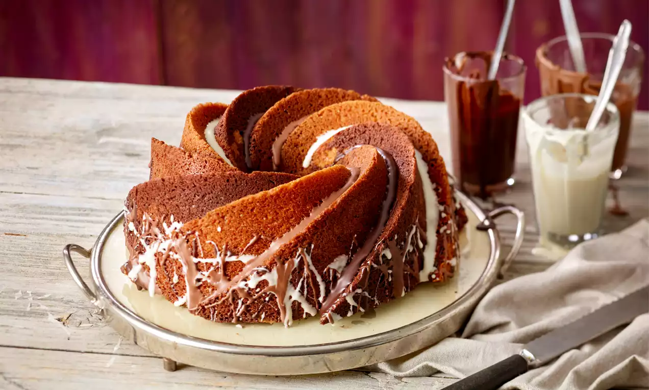 Gugelhupf aus drei Schokoladensorten mit marmoriertem Muster und weißer Glasur auf einer silbernen Platte, umgeben von Gläsern und Stofftuch
