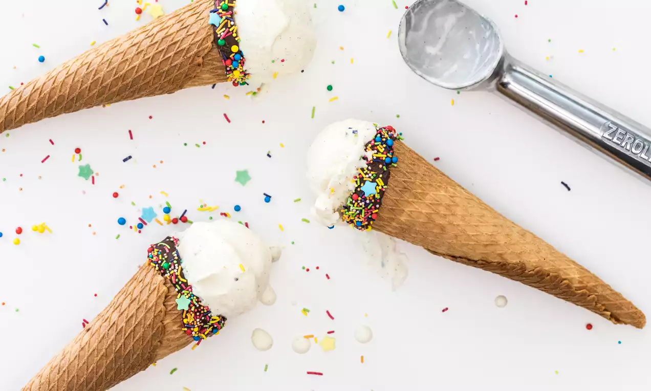 Conos de barquillo dorado con bordes decorados de chispas coloridas, rellenos de helado cremoso blanco sobre fondo con confites dispersos