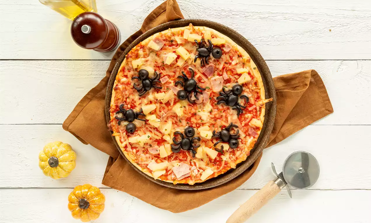 Pizza dorée saupoudrée de fromage et morceaux d’ananas avec des araignées en olives noires posée sur une serviette marron