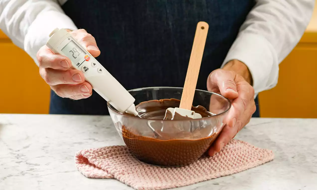 Glas Schüssel mit glänzend brauner, cremiger Kuvertüre auf rosa Stoffunterlage, Thermometer und Spatel werden von Händen gehalten