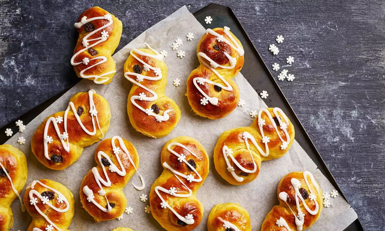 Gyllene lussekatter formade som S, dekorerade med vit kristyr och russin, placerade på bakplåtspapper med vita snöflingeströssel
