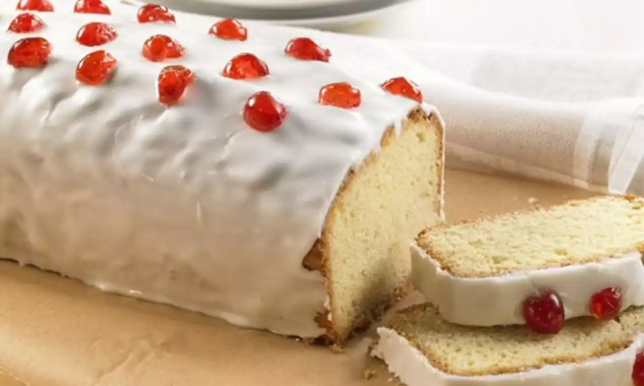 Ein länglicher Rührkuchen mit weißem Zuckerguss, dekoriert mit roten kandierten Kirschen, auf braunem Papier und mit angeschnittenen Scheiben