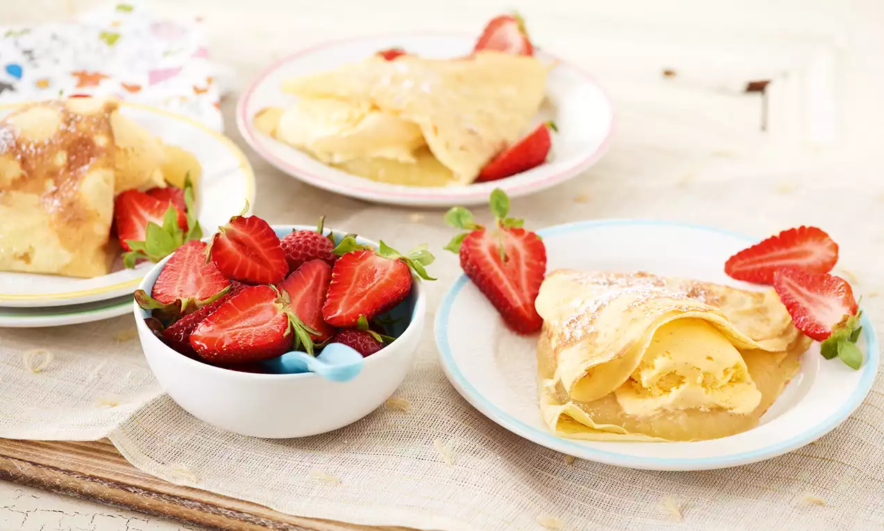 Goldgelbe Crêpes mit Apfelmusfüllung, zart bestäubt mit Puderzucker, dekoriert mit frischen roten Erdbeeren auf einem hellen Teller