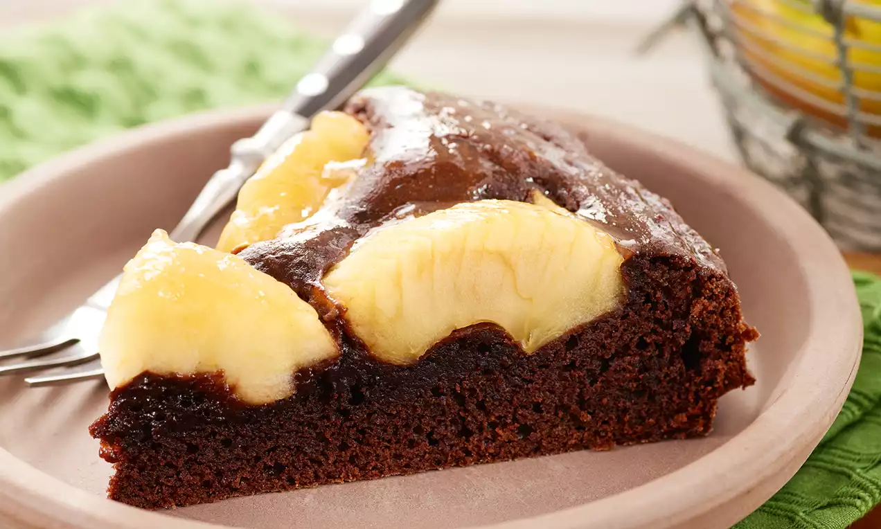 Fetta di torta al cioccolato morbida con mela dorata caramellata disposta a ventaglio su un piatto color crema accanto a una forchetta argento