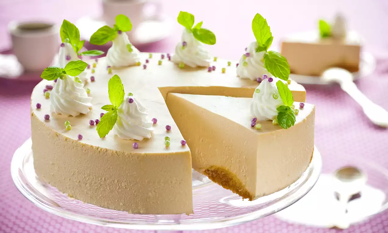 Okrugla cappuccino torta kremaste bež boje s podlogom od keksa, ukrašena bijelim šlagom, svježim listićima mente i sitnim šarenim perlicama