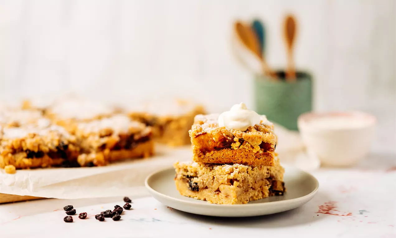 Два златисто-кафяви слоя ябълков крамбъл с натрошени маслени трохи, сервирани на светъл порцеланов съд и украсени с бяла сметана