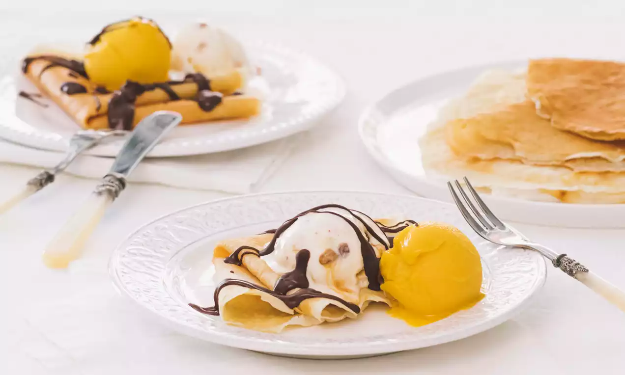 Crepas finas ligeramente doradas dobladas sobre helado cremoso de vainilla y mango, decoradas con hilos de chocolate en platos blancos