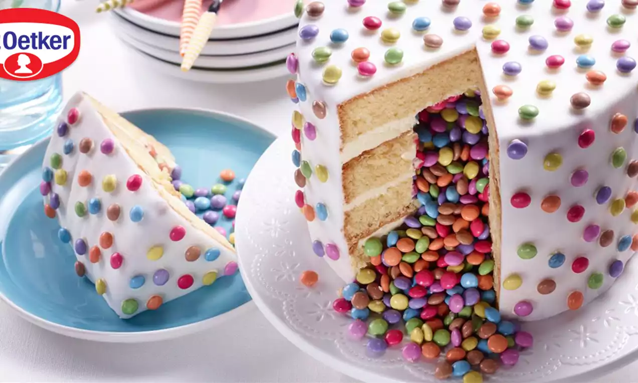 Witte fondanttaart met drie lagen biscuit, bedekt met kleurrijke chocoladeparels, gevuld met losse snoepjes en geserveerd op een wit bord