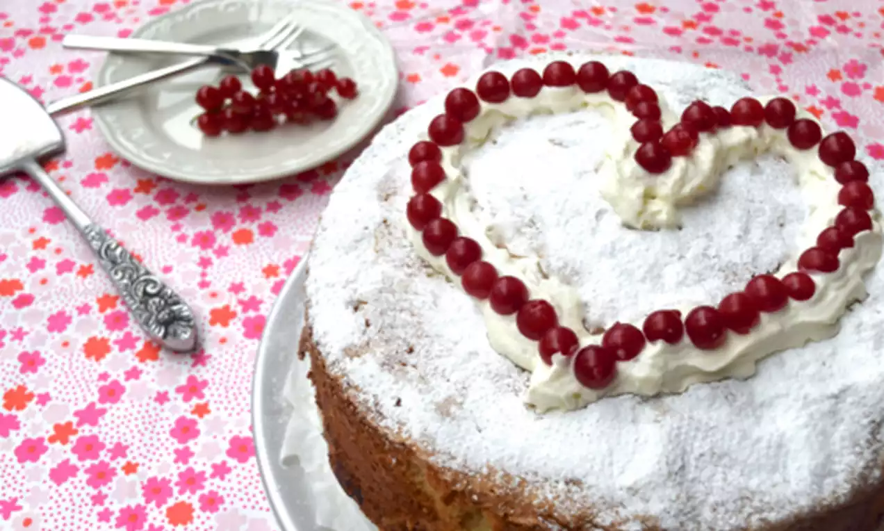 Ronde citroentaart met poedersuiker, slagroom en een hart van rode bessen, op een wit bord met een bloemenpatroon ondergrond