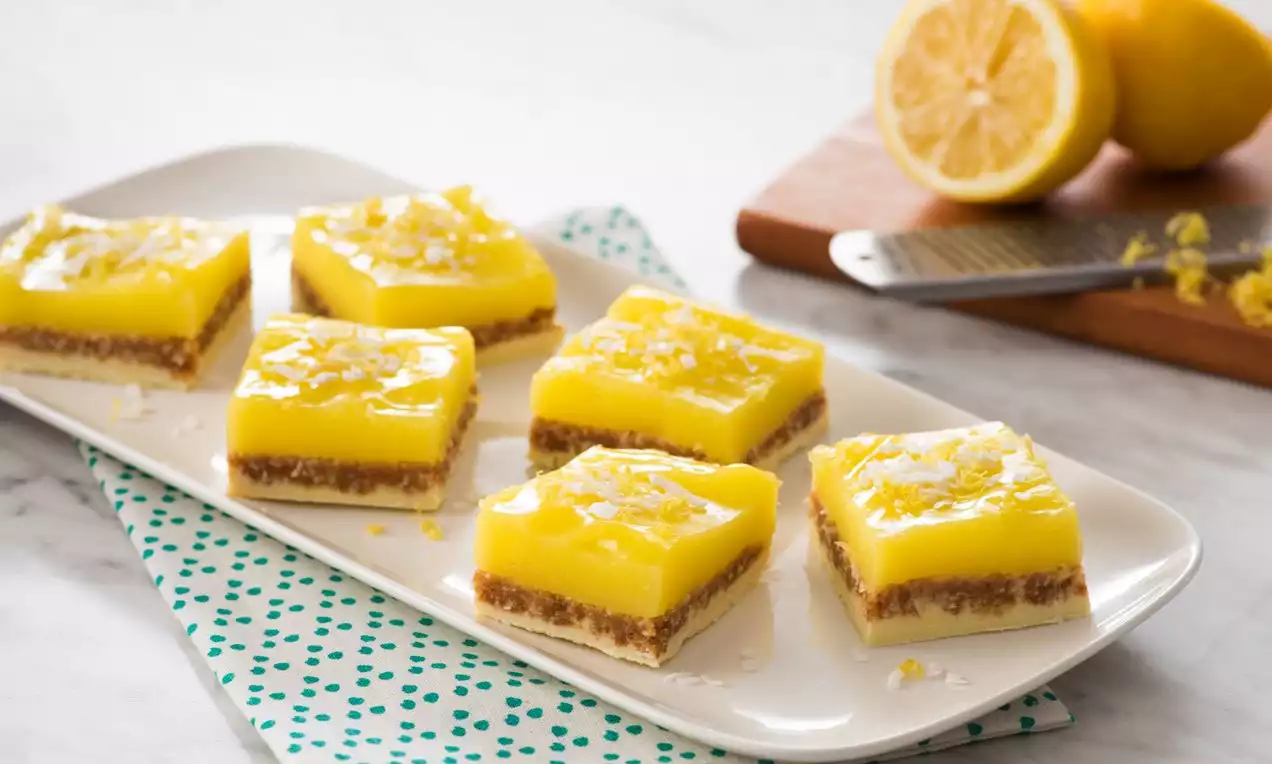 Carrés jaunes vifs au citron et lime avec une couche brune croustillante, parsemés de zestes d’agrumes sur un plat blanc rectangulaire