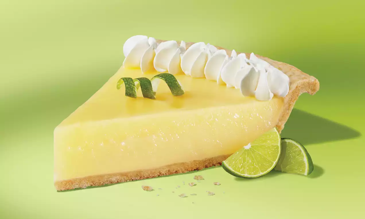 Key lime pie slice with smooth pale yellow filling, flaky crust, topped with whipped cream swirls, lime zest curls, and lime slices
