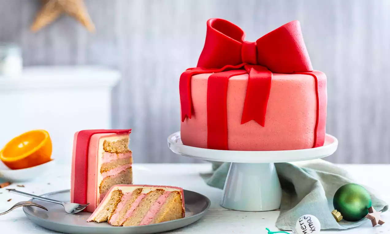 Julegavekake med rødt marsipanbånd og stor sløyfe på hvitt kakefat, ved siden av kakeskiver på grå tallerken med juletrekuler