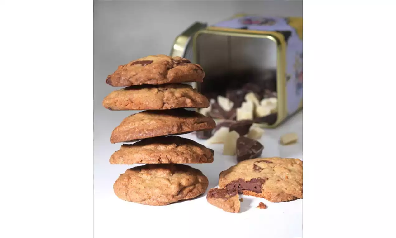 Fem gylne cookies med biter av mørk og hvit sjokolade stablet høyt foran en åpen metallboks med spredte sjokoladebiter rundt