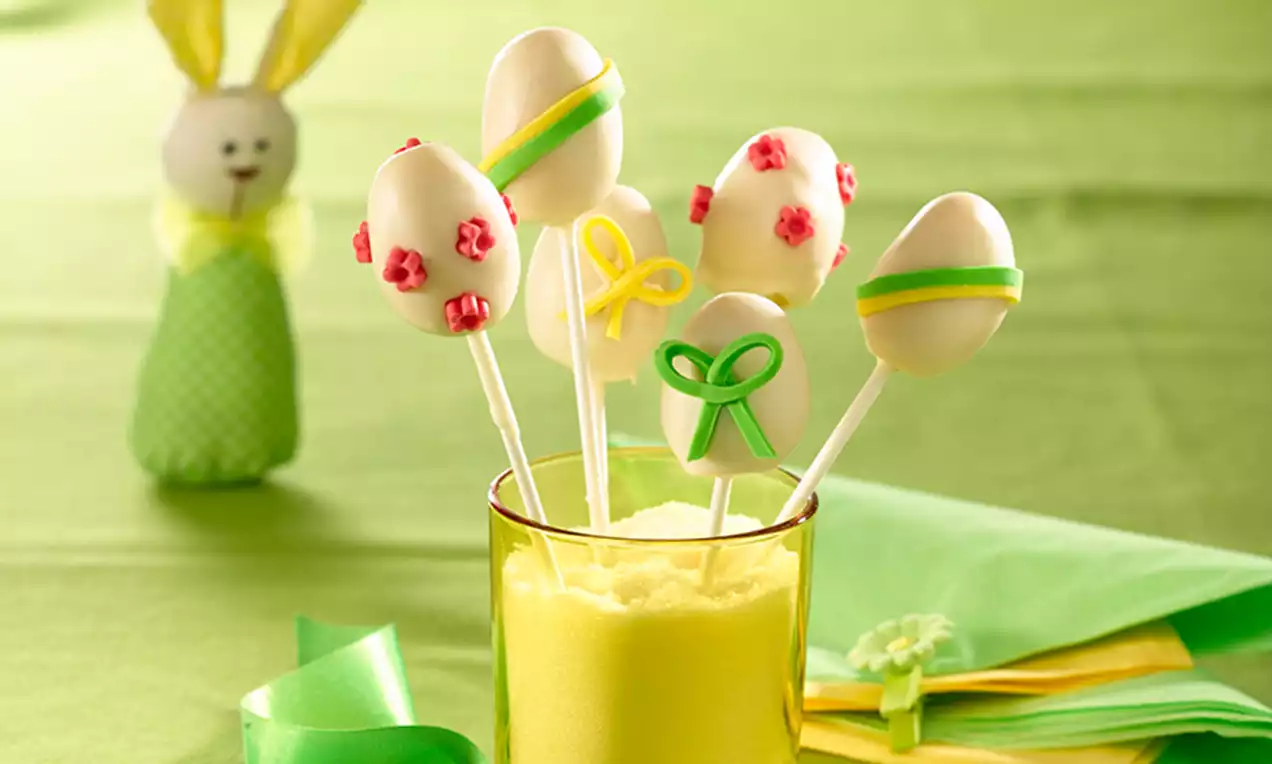 Cake pops en forme d’œufs de Pâques blancs décorés de rubans colorés et de fleurs roses, dressés dans un verre rempli de sucre jaune