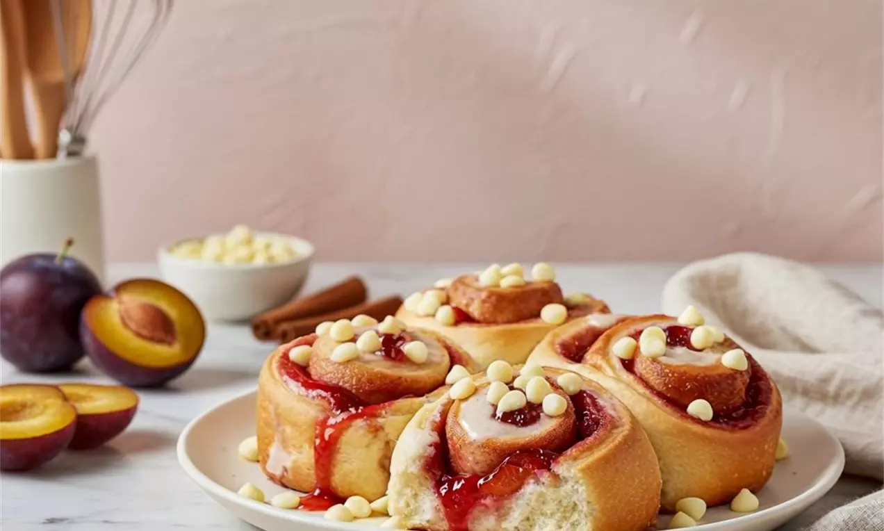 Picture - Golden vanilla rolls swirled with glossy plum filling, topped with creamy white chocolate drops on a white plate, surrounded by fresh plums and baking tools