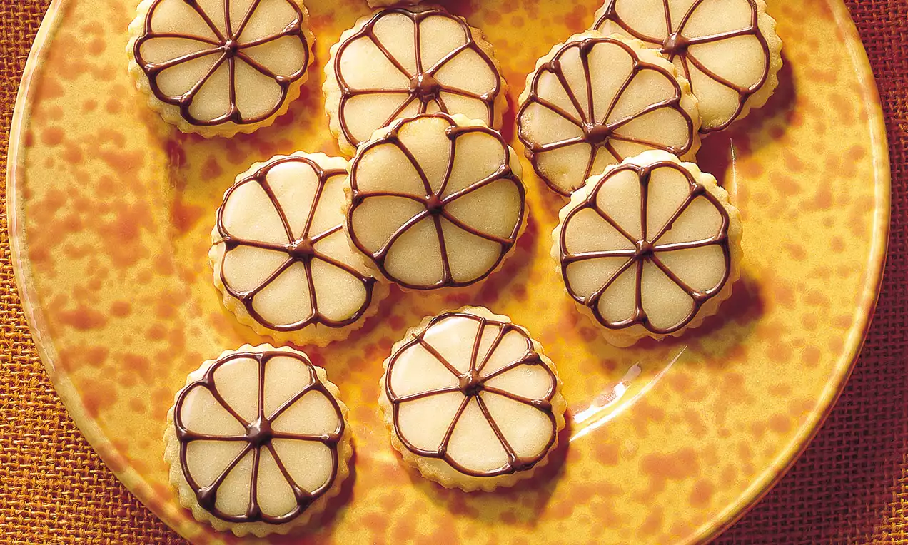 Biscotti rotondi dorati con glassa chiara e decorazioni al cioccolato a forma di petali serviti su un piatto giallo maculato