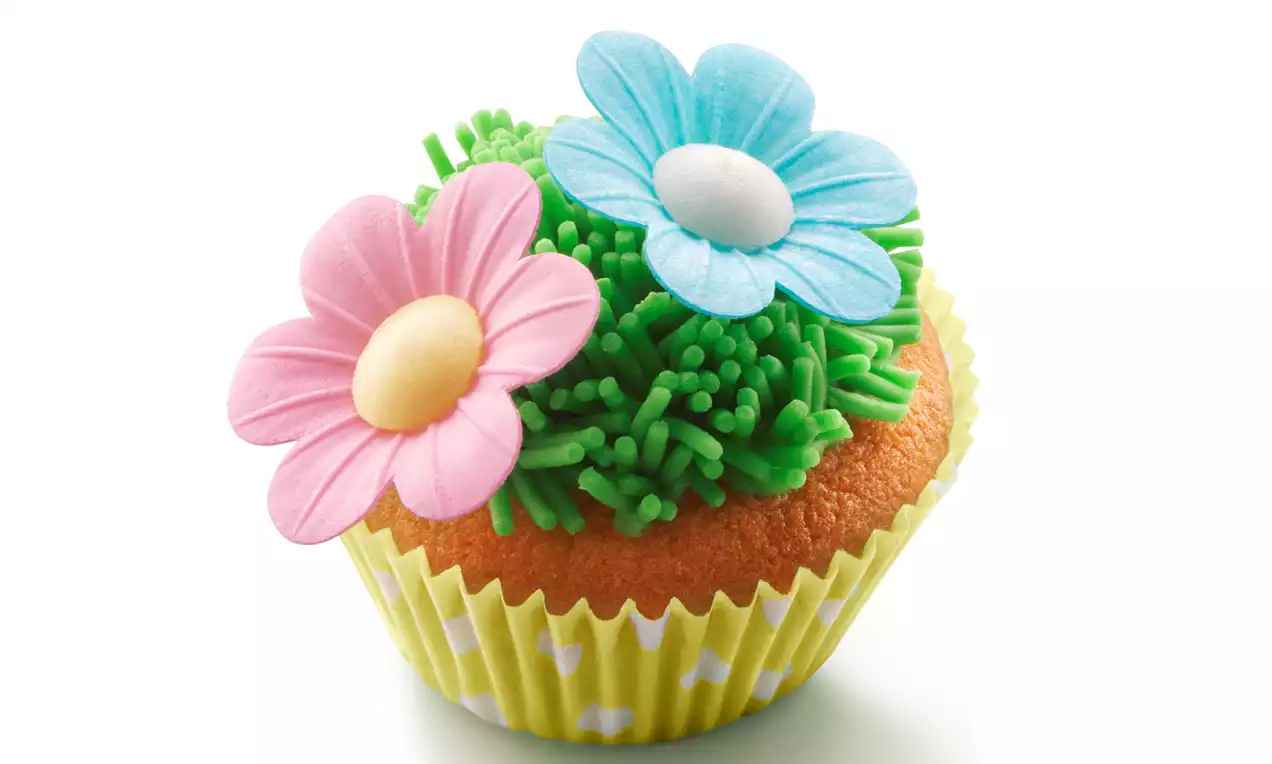 Cupcake in een geel bakingvormpje met groene grasachtige glazuurdecoratie en roze en blauwe suikerbloemen met witte harten