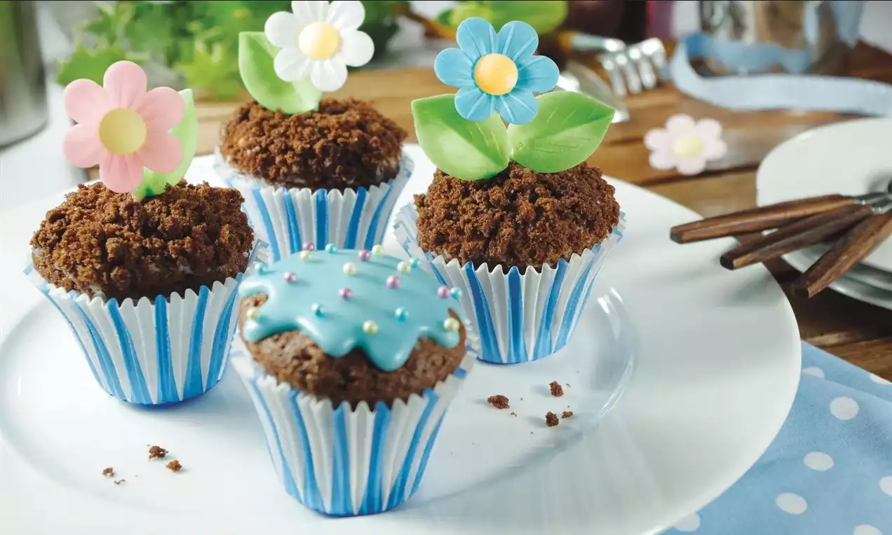 Čtyři čokoládové muffiny v modro-bílých košíčcích, zdobené barevnými jedlými květinami a posypané drobenkou, naaranžované na bílém talíři