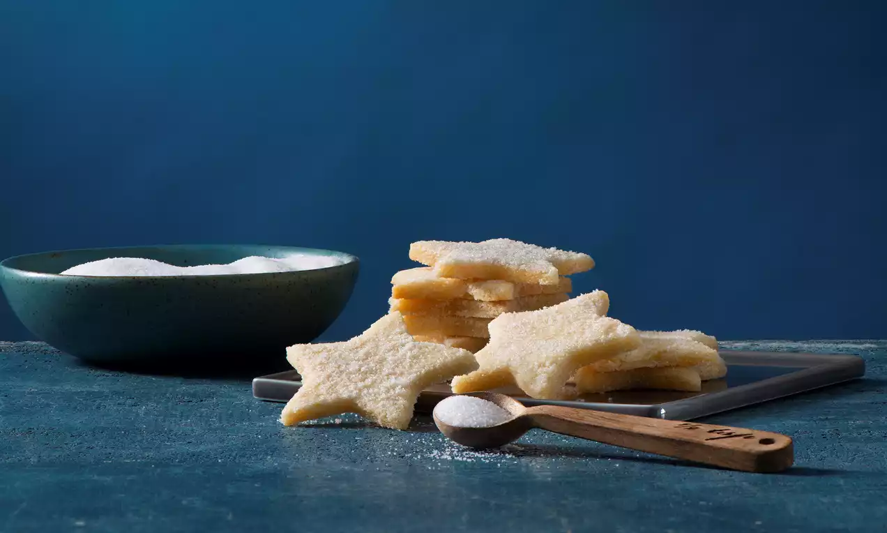 Golden vanilla sugar cookies shaped like five-pointed stars, stacked on a tray with a wooden spoon and a green bowl of sugar nearby
