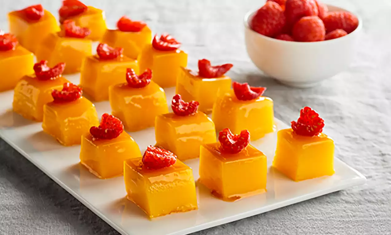 Cubos de gelatina amarilla brillante con textura suave, coronados por frambuesas partidas sobre una bandeja blanca junto a un cuenco de frambuesas